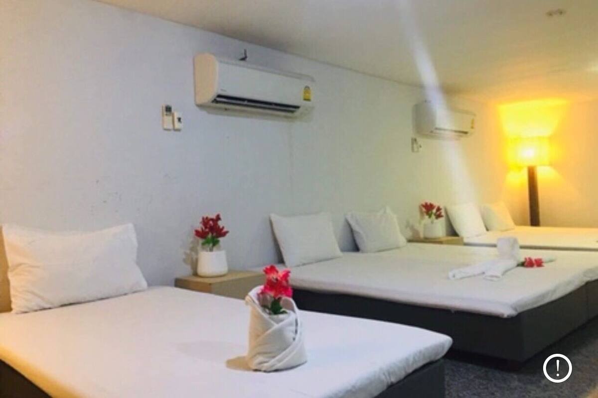 Two single beds with white linens, a wall-mounted air conditioner, a bedside table with a vase of flowers, and a lamp on the right side.
