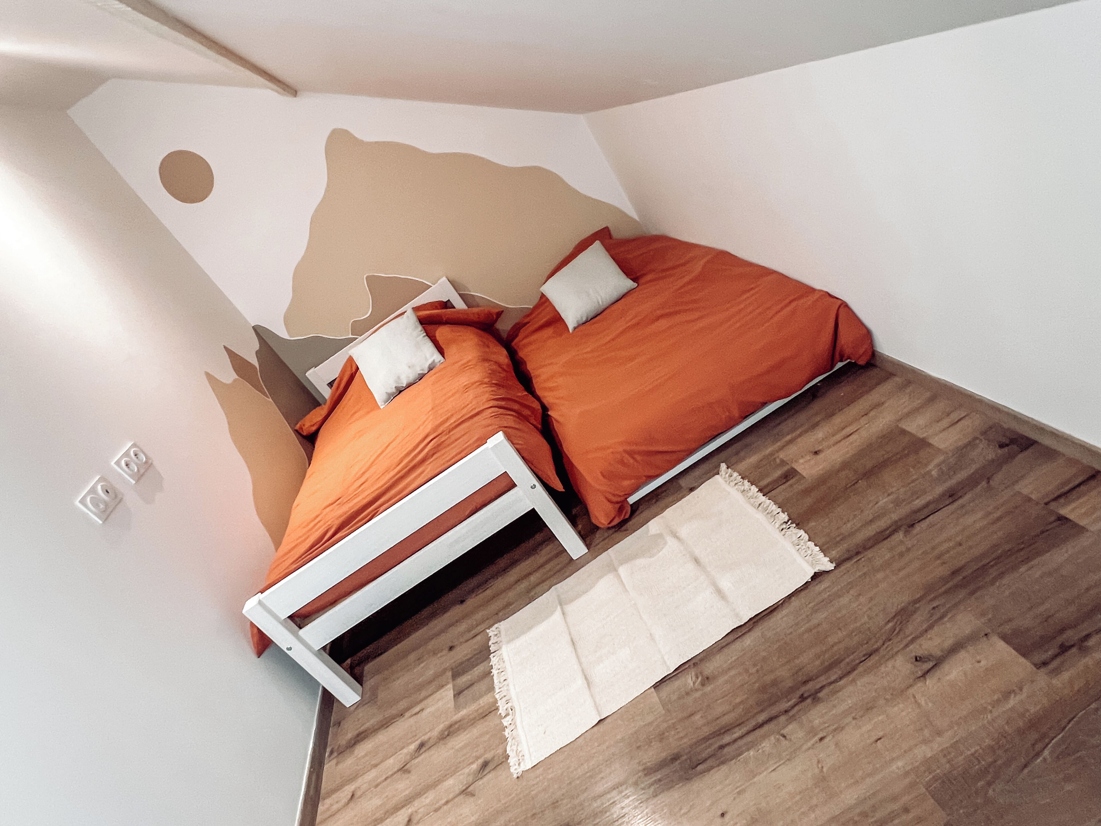 A bedroom with a white bed frame, two orange bedsheets, a white pillow, and a wooden floor.