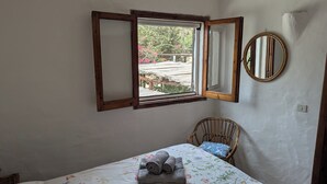 A bedroom with a bed, a window with a wooden frame, a wicker chair, and a round mirror on the wall. - Villa a few steps from the sea of Villasimius (Villasimius)
