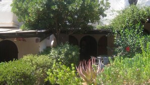 A courtyard with a large tree, a white building with blue accents, and a small balcony. - Villa a few steps from the sea of Villasimius (Villasimius)