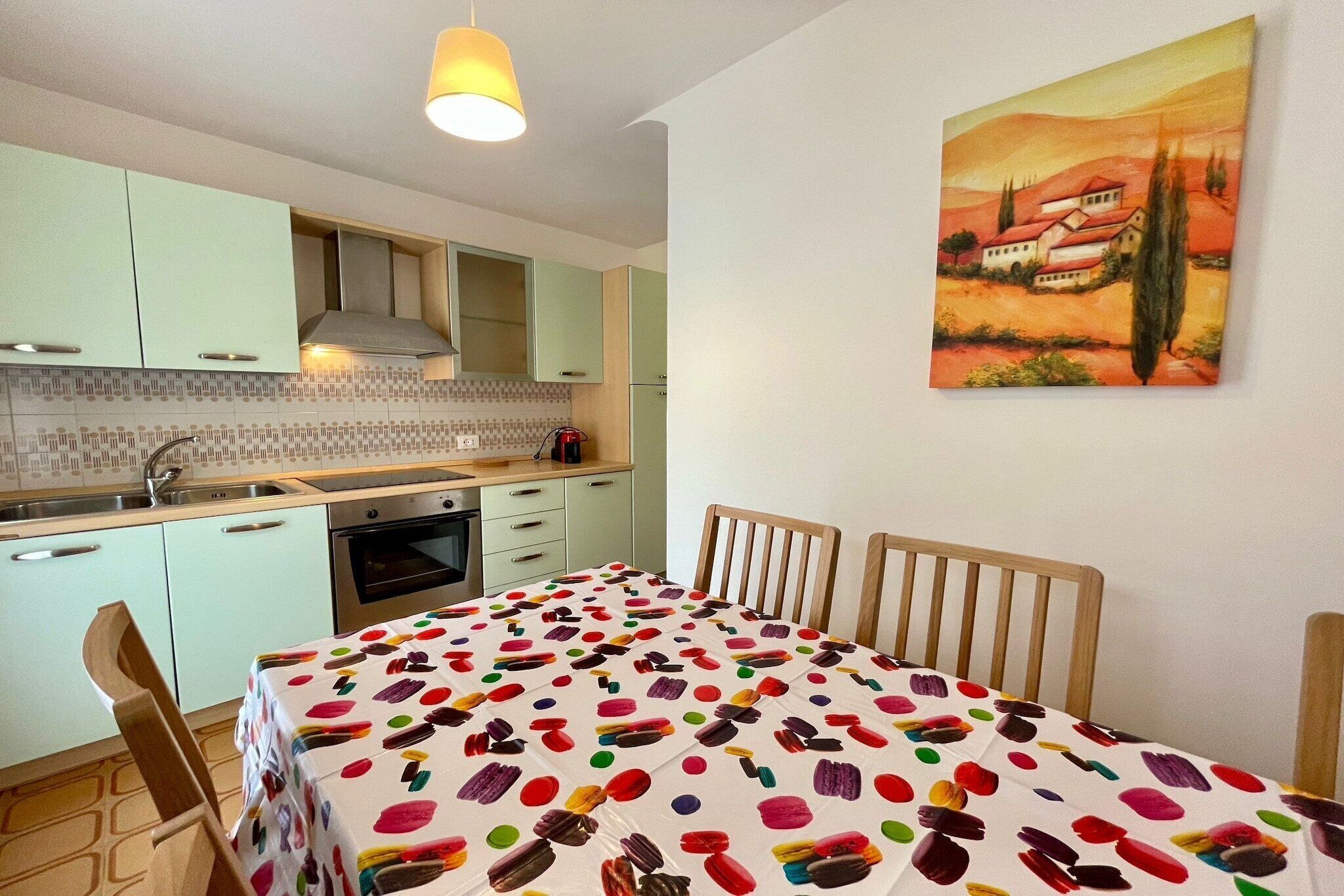 A kitchen with light green cabinets, a tiled backsplash, and a dining table with a colorful tablecloth.