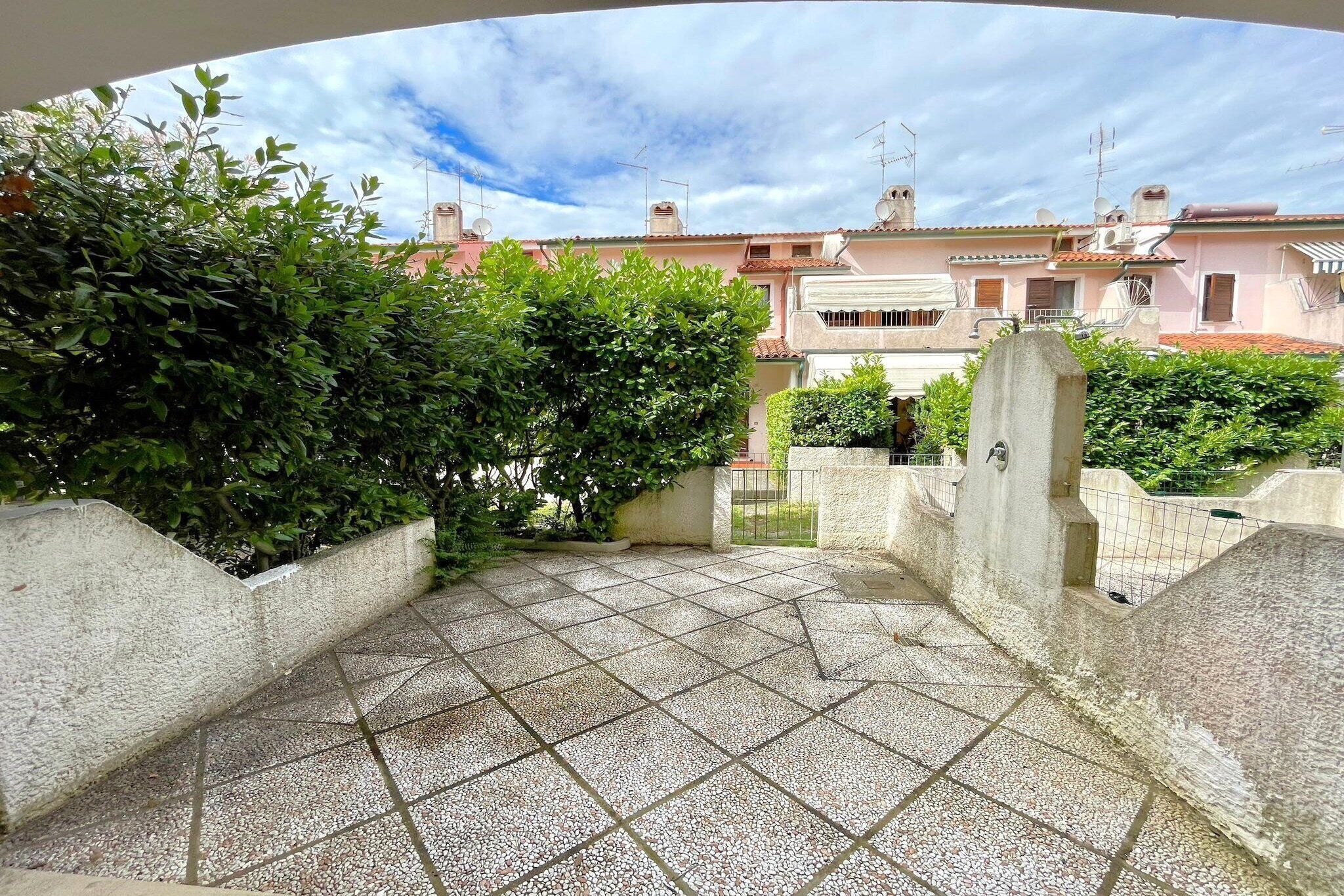 A rooftop terrace with tiled flooring, surrounded by greenery and buildings in the background.
