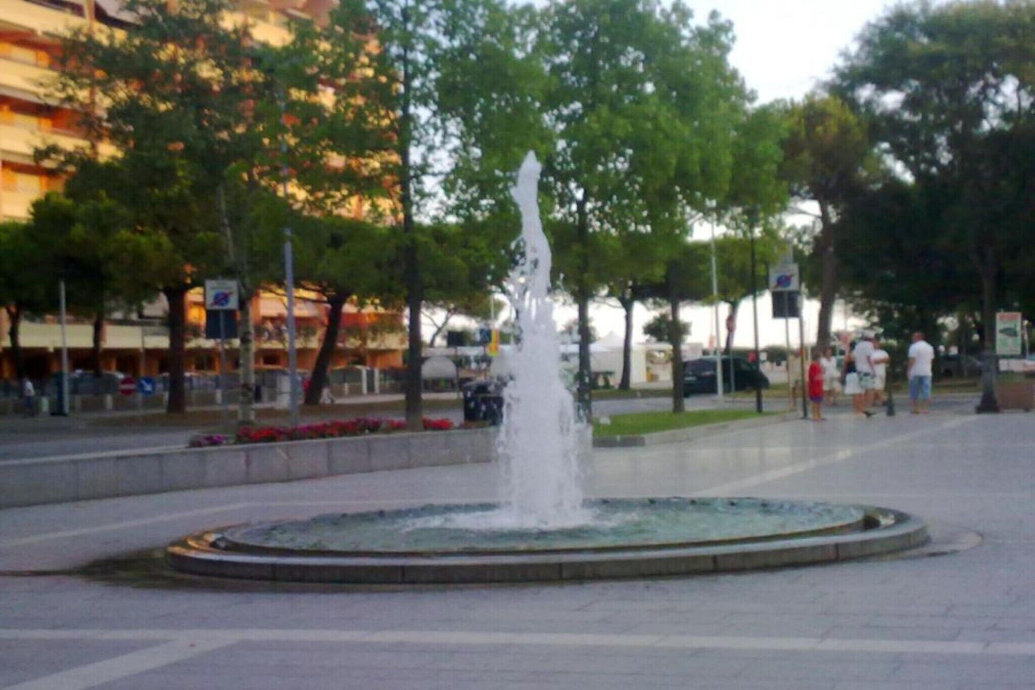 A public fountain with water jets in a plaza surrounded by trees and buildings.