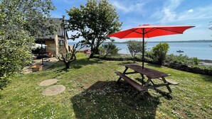 Un patio con mesa de picnic y una sombrilla roja, con vista a un lago con barcos.