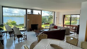 Amplia sala de estar con un gran ventanal con vistas a un lago, amueblada con un sofá, sillas y una mesa de comedor.