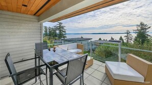 A modern outdoor patio with a glass table and chairs, a cushioned seating area, and a view of the water and trees.