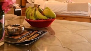 Una habitación de hotel con una cama, una mesita de noche con un cuenco de plátanos, una revista y un jarrón de flores.