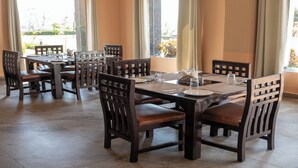 Un comedor con varias mesas y sillas de madera, ventanas amplias con cortinas y piso de baldosas.