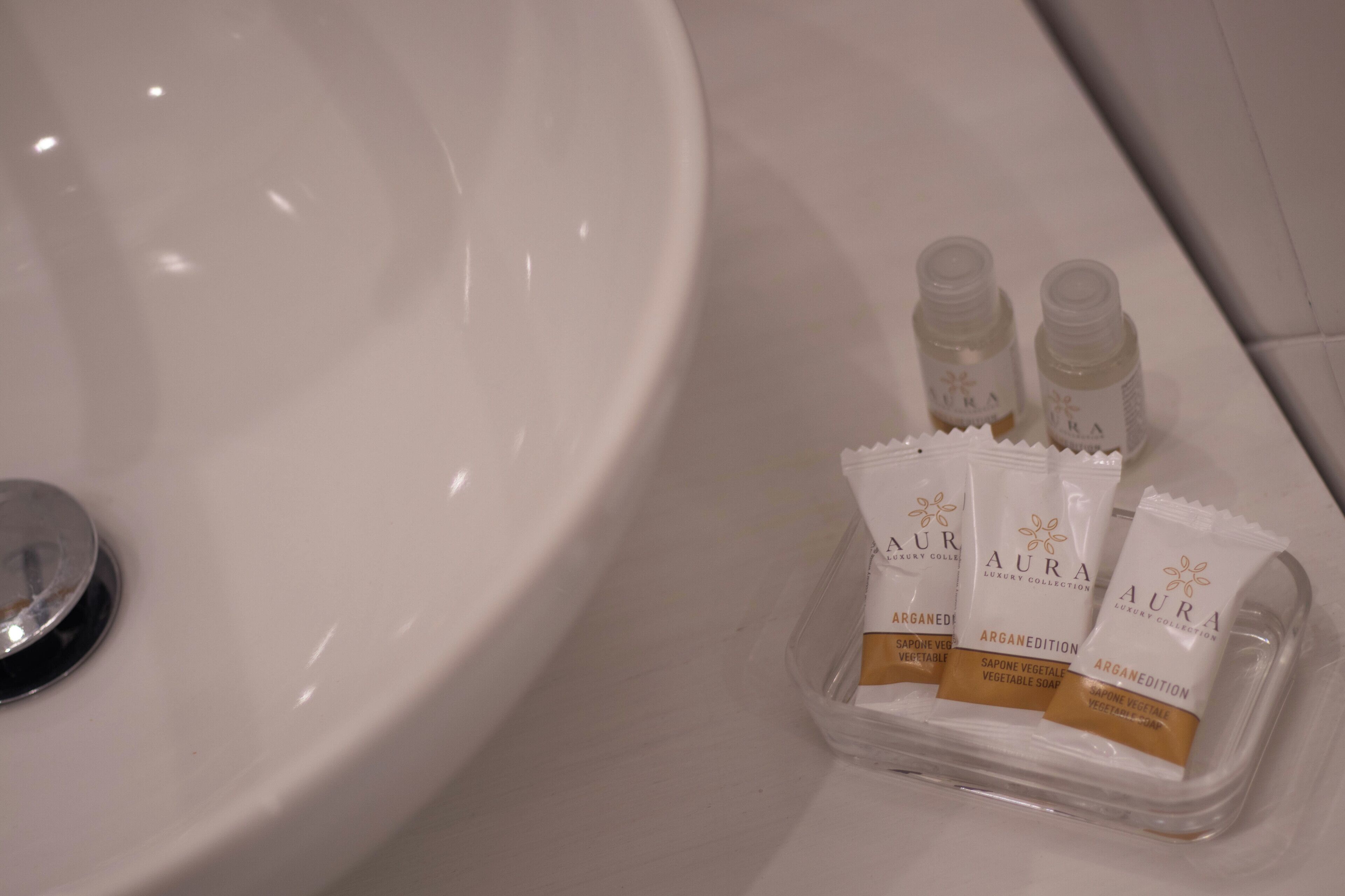 Un évier blanc, à côté duquel sont placés trois flacons de savon à l’huile d’argan de la marque « AURA ».