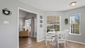Un coin repas avec une table ronde et des chaises blanches, une horloge au mur et une fenêtre avec des stores.