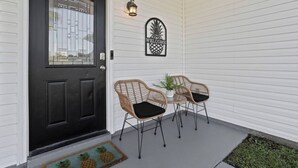 Une porte d’entrée noire avec un panneau de verre, deux chaises en osier avec des coussins noirs, une petite table sur laquelle est posée une plante en pot, et une pancarte d’accueil ornée d’un motif représentant un ananas.