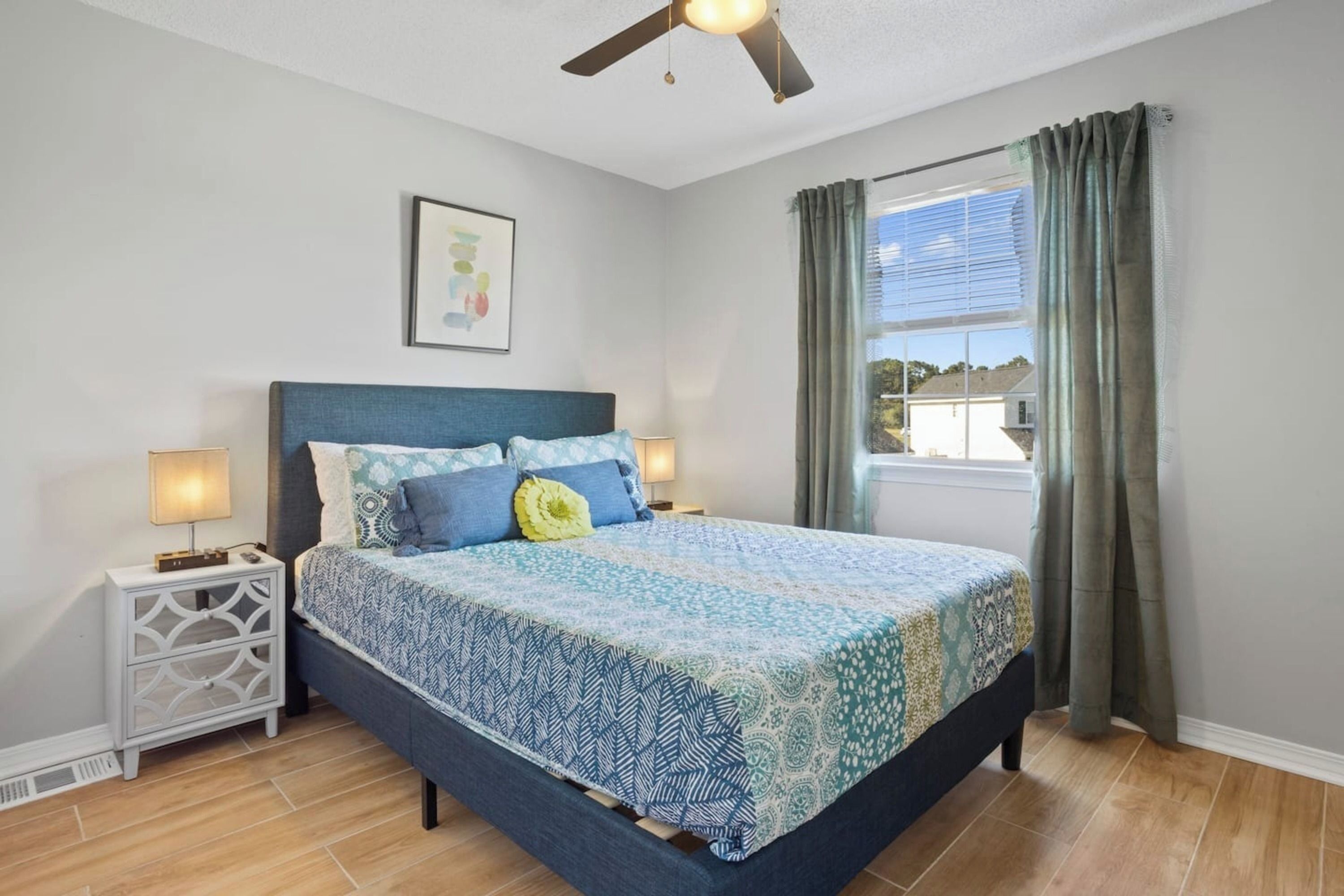 A bedroom with a bed, nightstands, a ceiling fan, a window with curtains, and a wall-mounted picture.