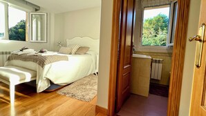 A bedroom with a bed, bedside table, and a window. There is a wooden door leading to a bathroom with a sink and a window. - Casa Montañesa al Lado de la Playa (Somo)