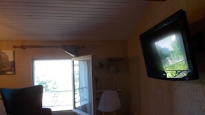 A room with a television mounted on the wall, a chair, a small ottoman, and a window with a view of greenery outside. - Tramontane, bedroom, and TV room. Near Avignon. (Jonquerettes)
