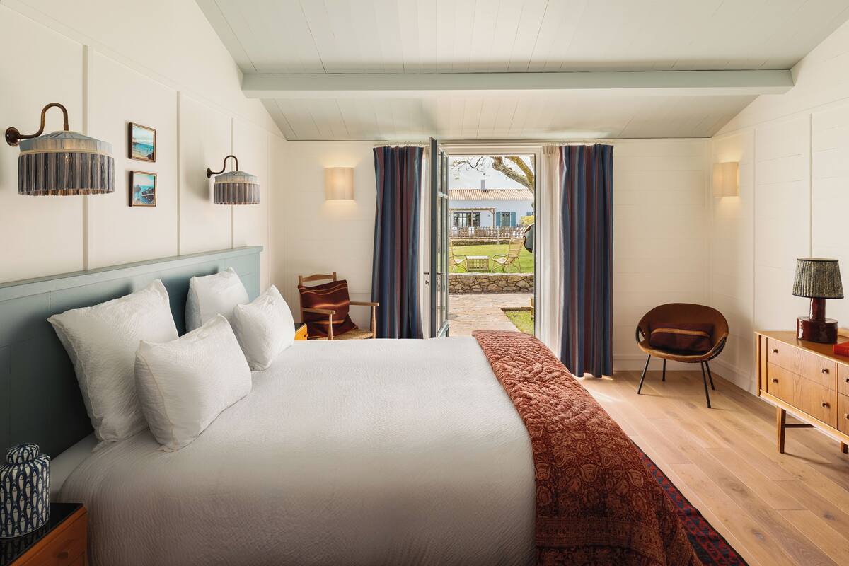 Un dormitorio con una cama grande, un aparador de madera, una silla y vistas al exterior a través de un ventanal.