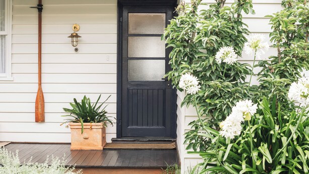 A black door with a glass panel, a wooden porch, a potted plant, and a wall-mounted light fixture. - Benambra Cottage (Queenscliff)