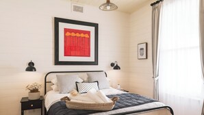 A neatly arranged bedroom with a bed, bedside tables, a framed artwork, and a window with curtains. - Benambra Cottage (Queenscliff)