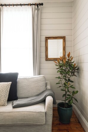 A room with a light-colored sofa, a potted plant, a mirror, and a window with curtains. - Benambra Cottage (Queenscliff)