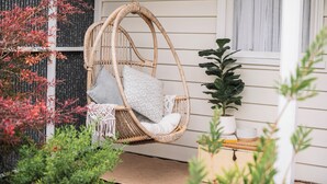 A wicker hanging chair with cushions, a potted plant, and a small table with a book and a candle. - Benambra Cottage (Queenscliff)