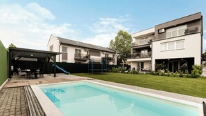 Una piscina con un porche cubierto, un césped y edificios de apartamentos modernos al fondo.