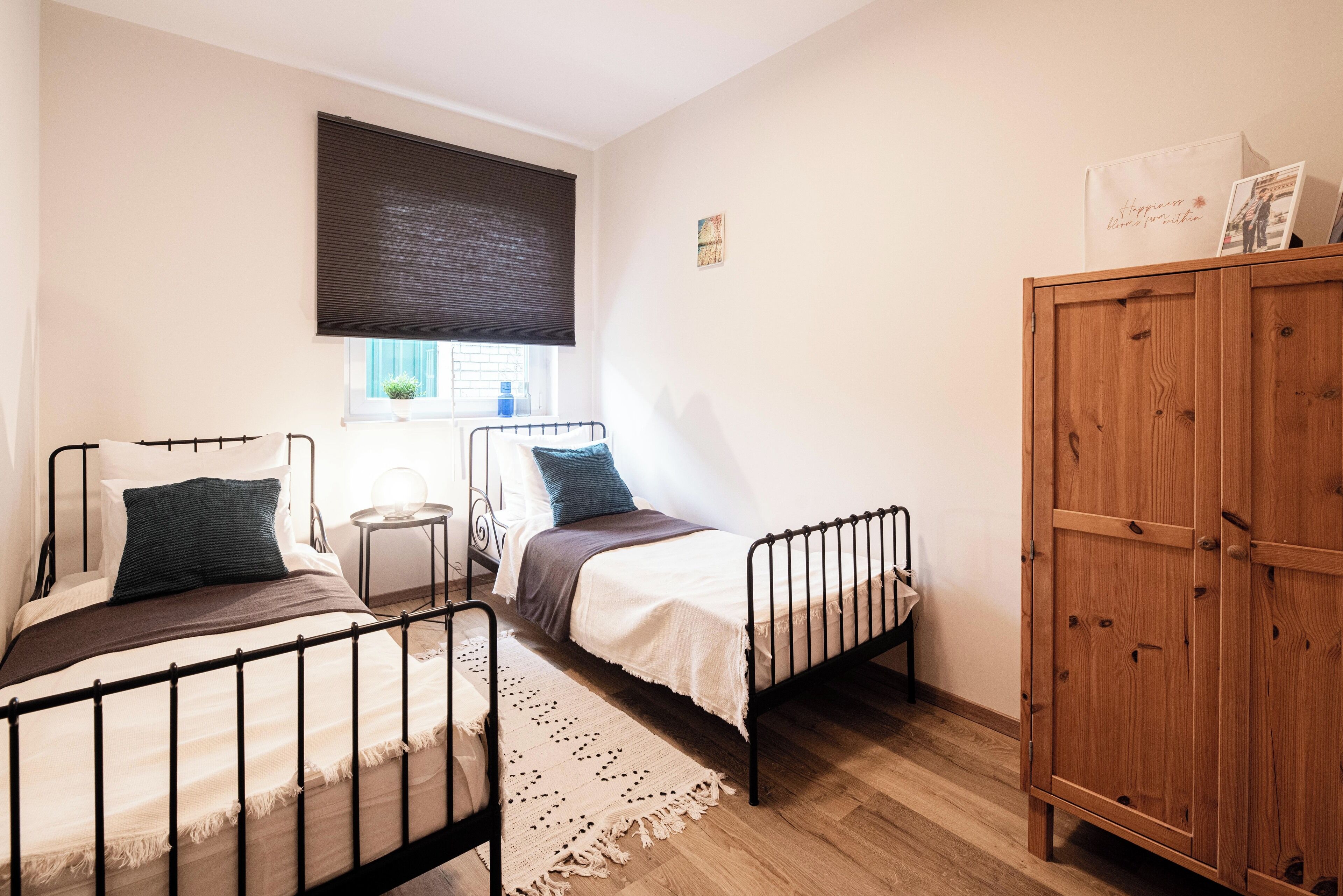 A bedroom with two metal beds, a wooden wardrobe, a small bedside table, a window with a blind, and a wall-mounted artwork.