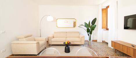 Un salon moderne avec un canapé beige, une table basse, un lampadaire, une grande plante en pot et un téléviseur fixé au mur.