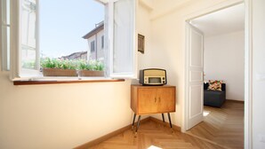 A room with a wooden floor, a window with a view of buildings and flowers, a small wooden cabinet with a radio, and a door leading to another room. - Singer House in Center Town (Lucca)