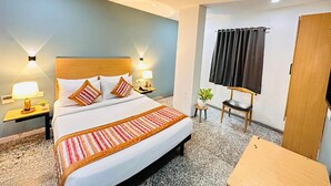 A hotel room with a large bed, a ceiling fan, two bedside lamps, a small table, and a window with curtains. - Qotel Comfort Golden Saffron Punjabi Bagh (New Delhi)