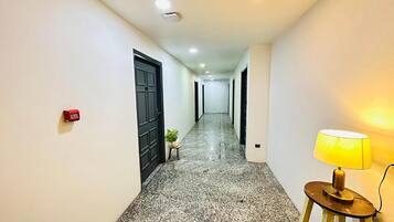 A hallway with a granite floor, white walls, and a black door. There is a small table with a plant and a lamp on the right.