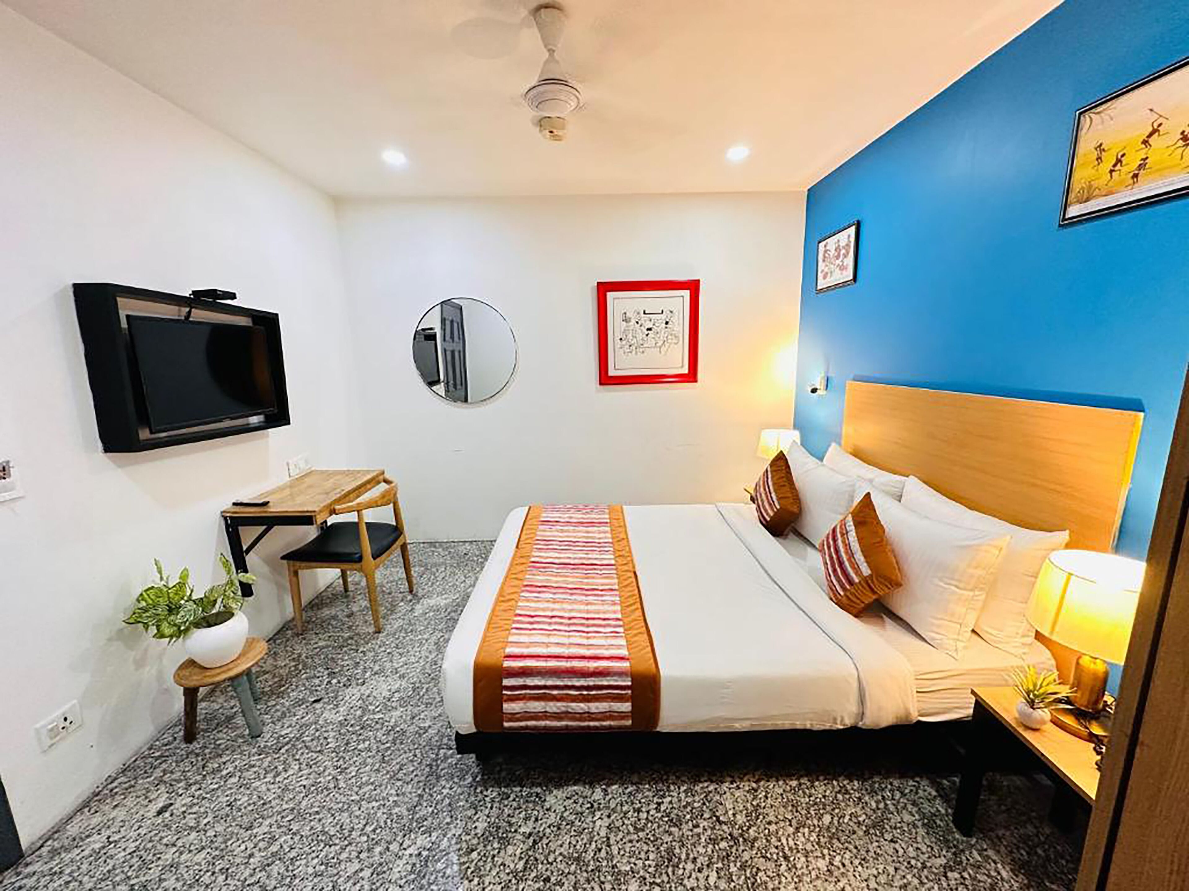 A hotel room with a bed, bedside table, lamp, TV, desk, chair, and a small plant.