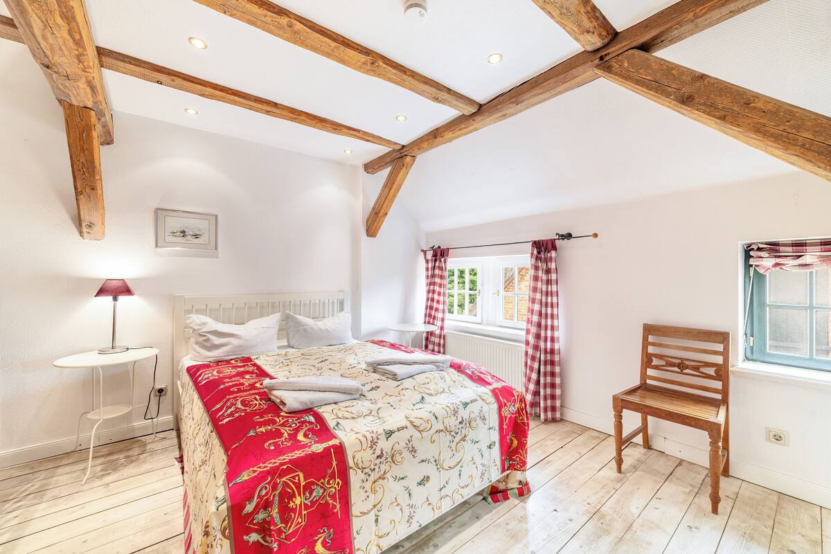 A bedroom with a wooden ceiling, a bed with a red and white patterned bedspread, a wooden chair, and a window with red curtains.