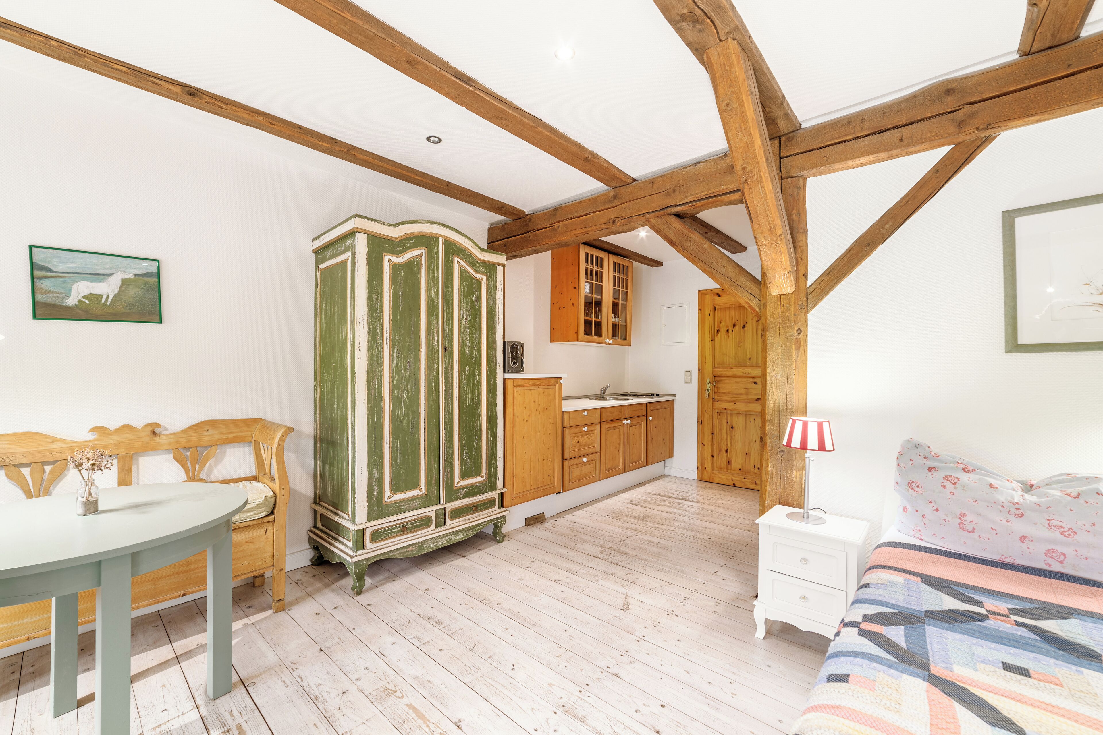 A bedroom with a wooden ceiling, a green wardrobe, a wooden cabinet, a white bedside table, and a bed with a floral-patterned bedspread.