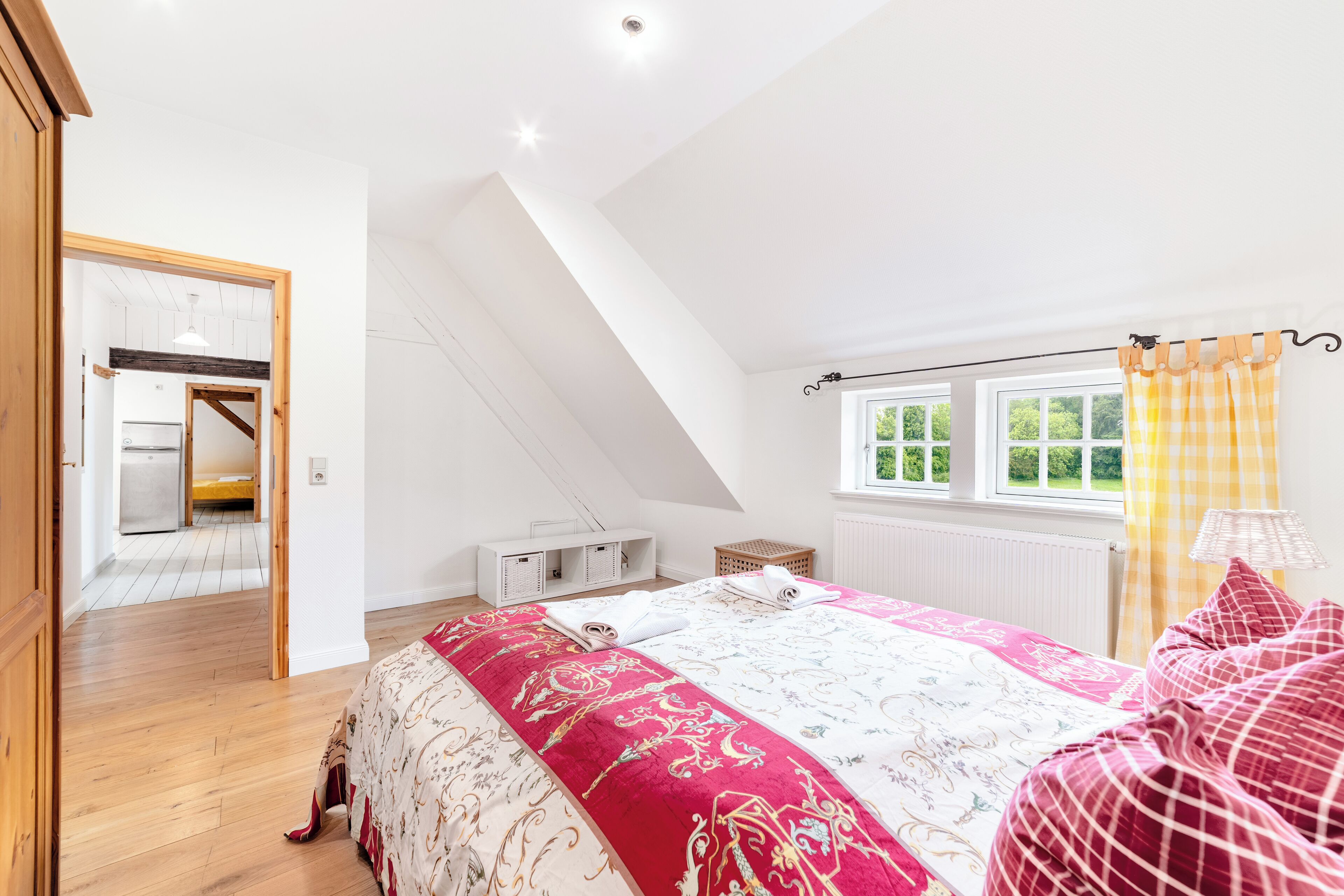 A bedroom with a sloped ceiling, a bed with patterned bedding, a window with yellow curtains, and a view of a kitchen and bathroom.