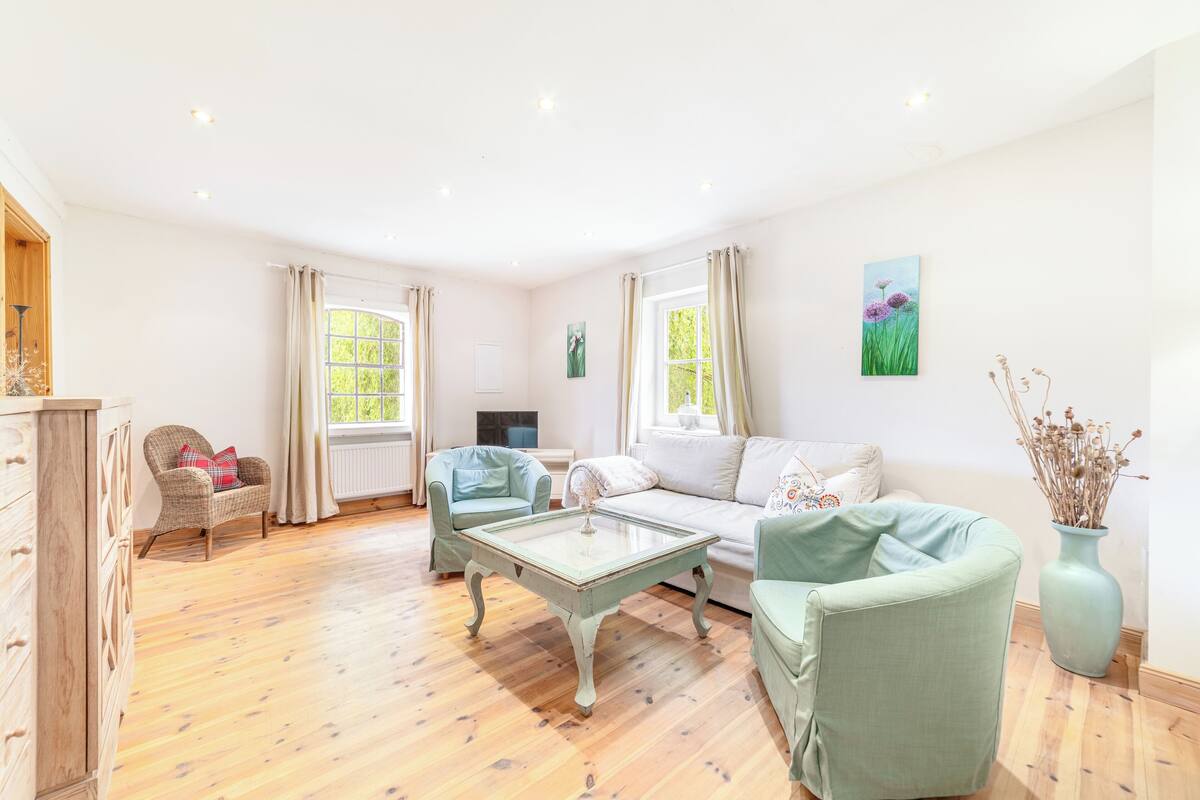 A spacious living room with a wooden floor, a white sofa, two armchairs, a glass coffee table, and a wooden cabinet.
