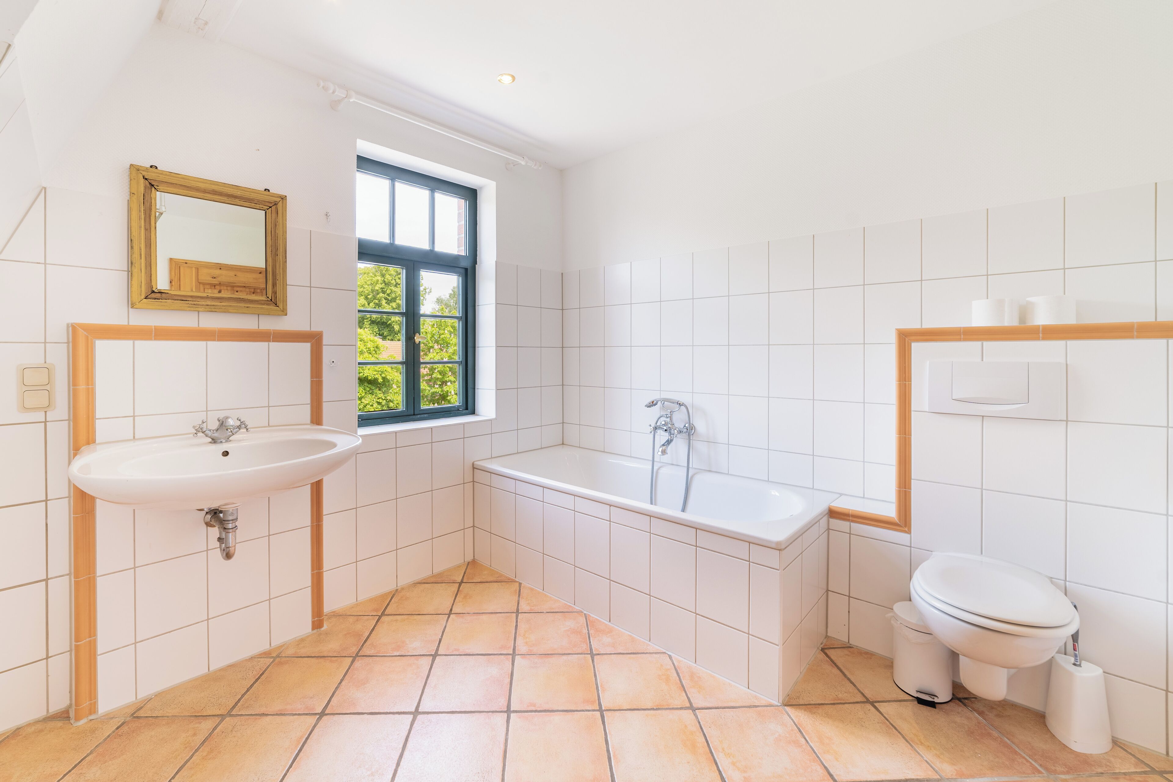 A bathroom with a sink, bathtub, and toilet, tiled in white, with a window providing natural light.