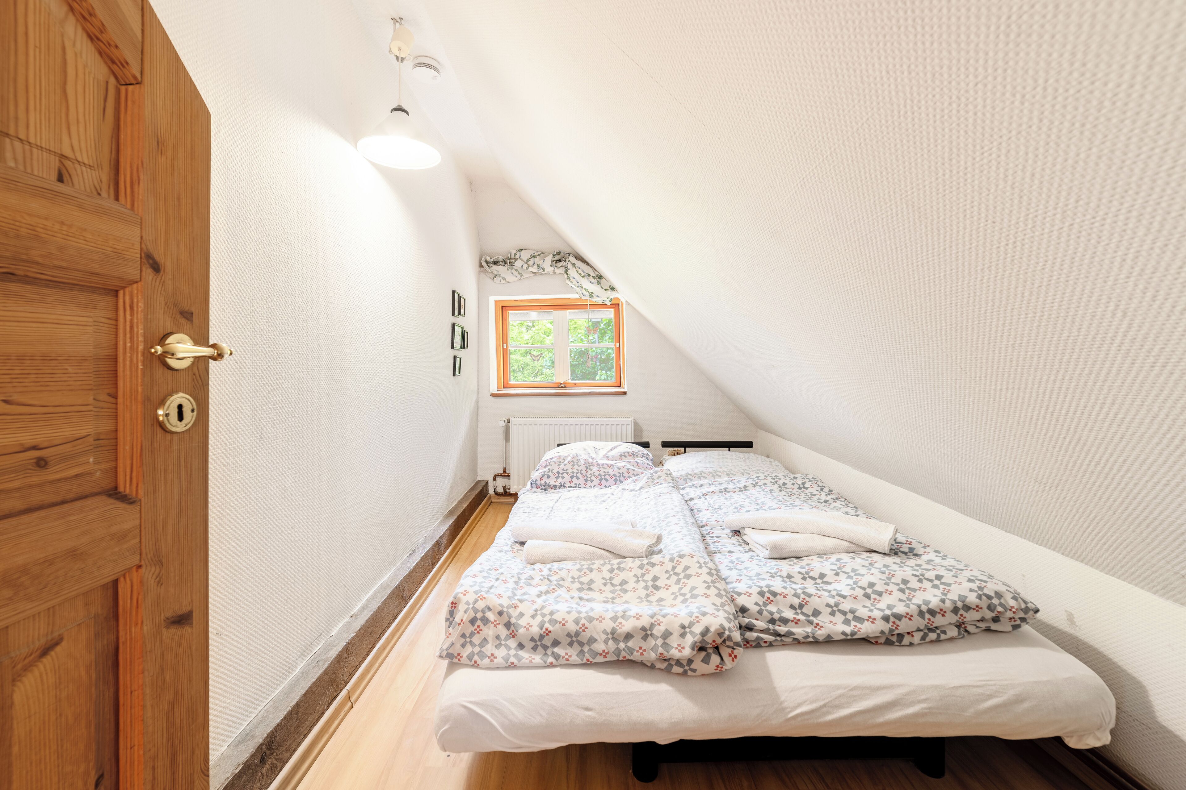 Una habitación pequeña y acogedora con techo inclinado, puerta de madera, ventana con vistas a la vegetación y cama con ropa de cama estampada.