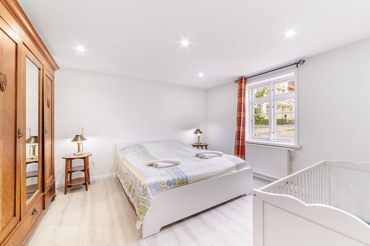 A bedroom with a bed, a wooden wardrobe, a small table with a lamp, a window with curtains, and a white crib.
