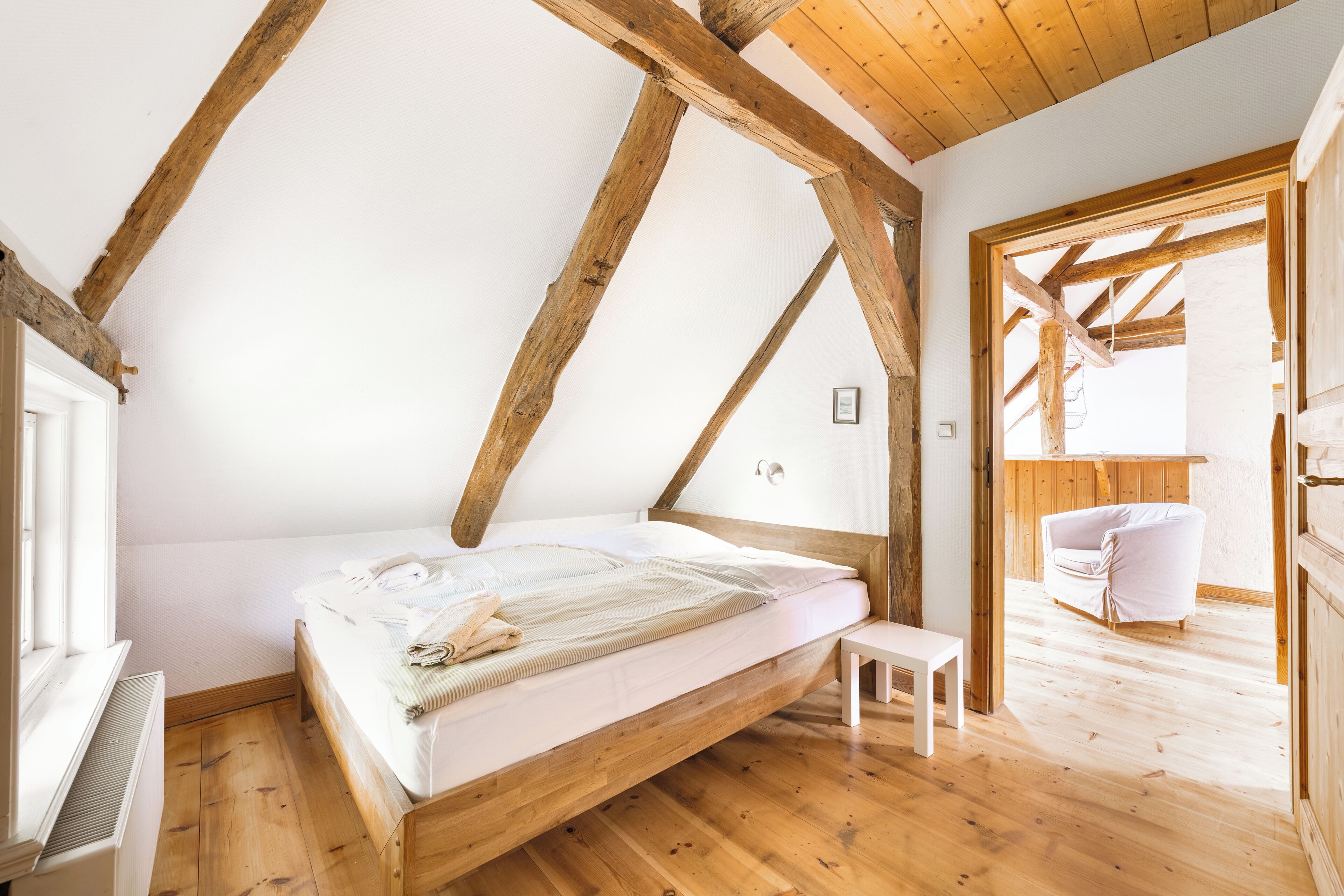 A bedroom with a sloped ceiling, wooden beams, a bed, a small table, and a view of a balcony.
