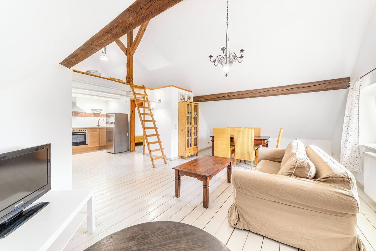 A spacious living area with a wooden ceiling, a sofa, a TV, a wooden table, and a kitchen in the background.