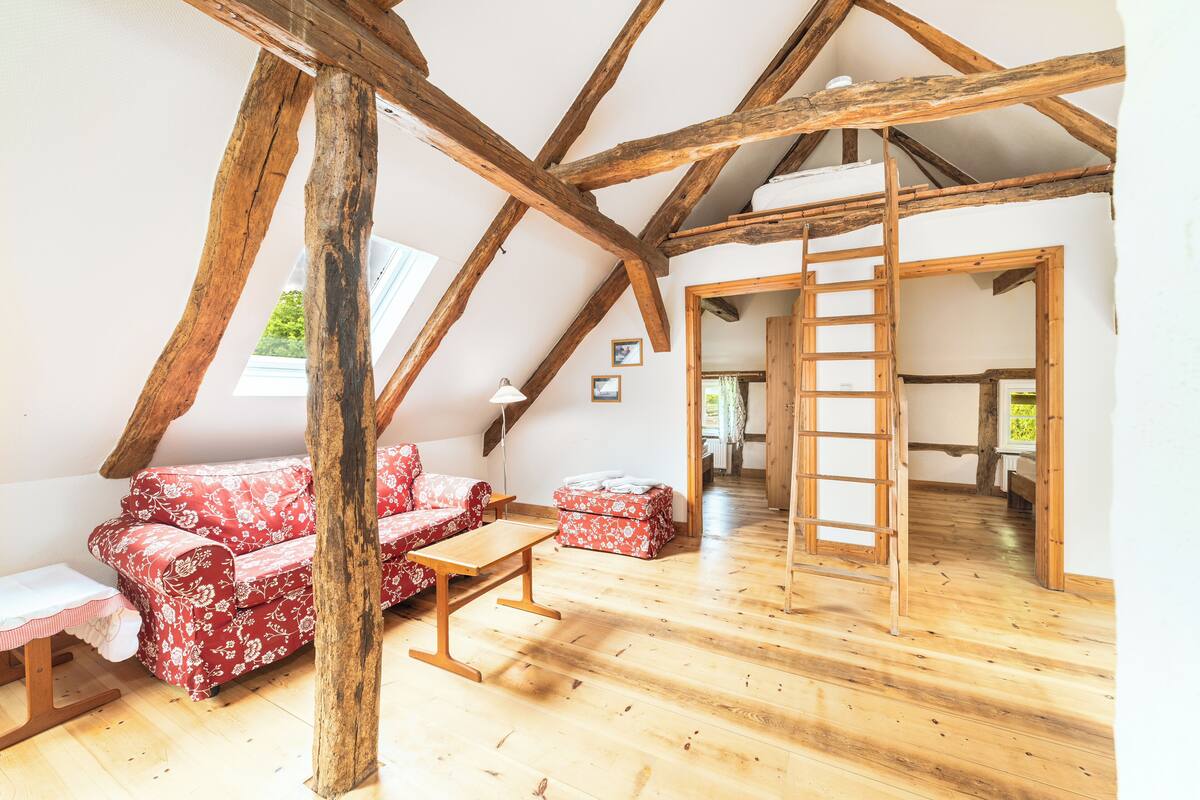 A loft-style room with exposed wooden beams, a red floral sofa, a wooden table, and a ladder leading to an upper level.