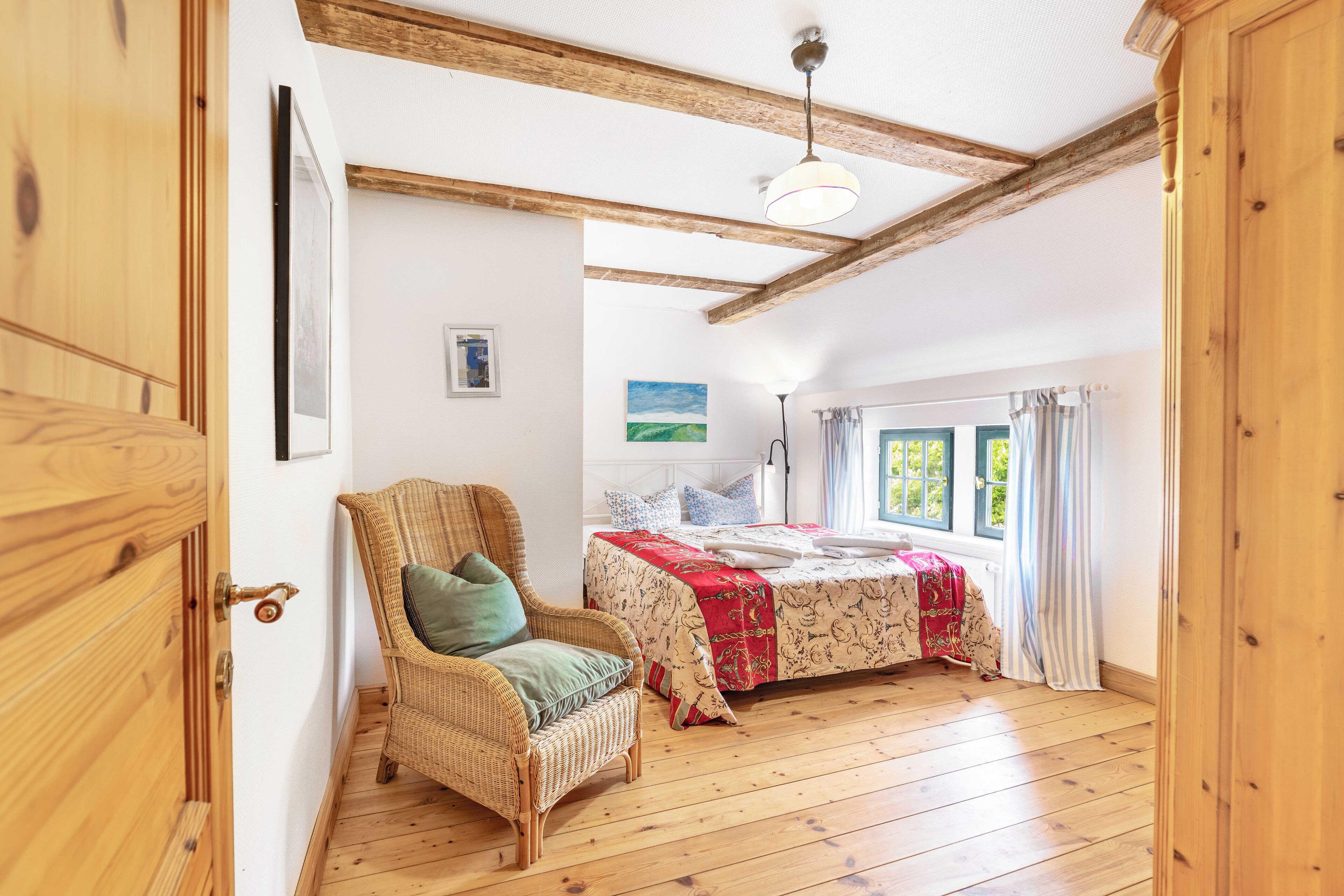 Ein Zimmer mit Holzboden, einem Bett mit rot-weißer Bettdecke, einem Korbstuhl mit grüner Sitzkissen und einem Bild an der Wand.