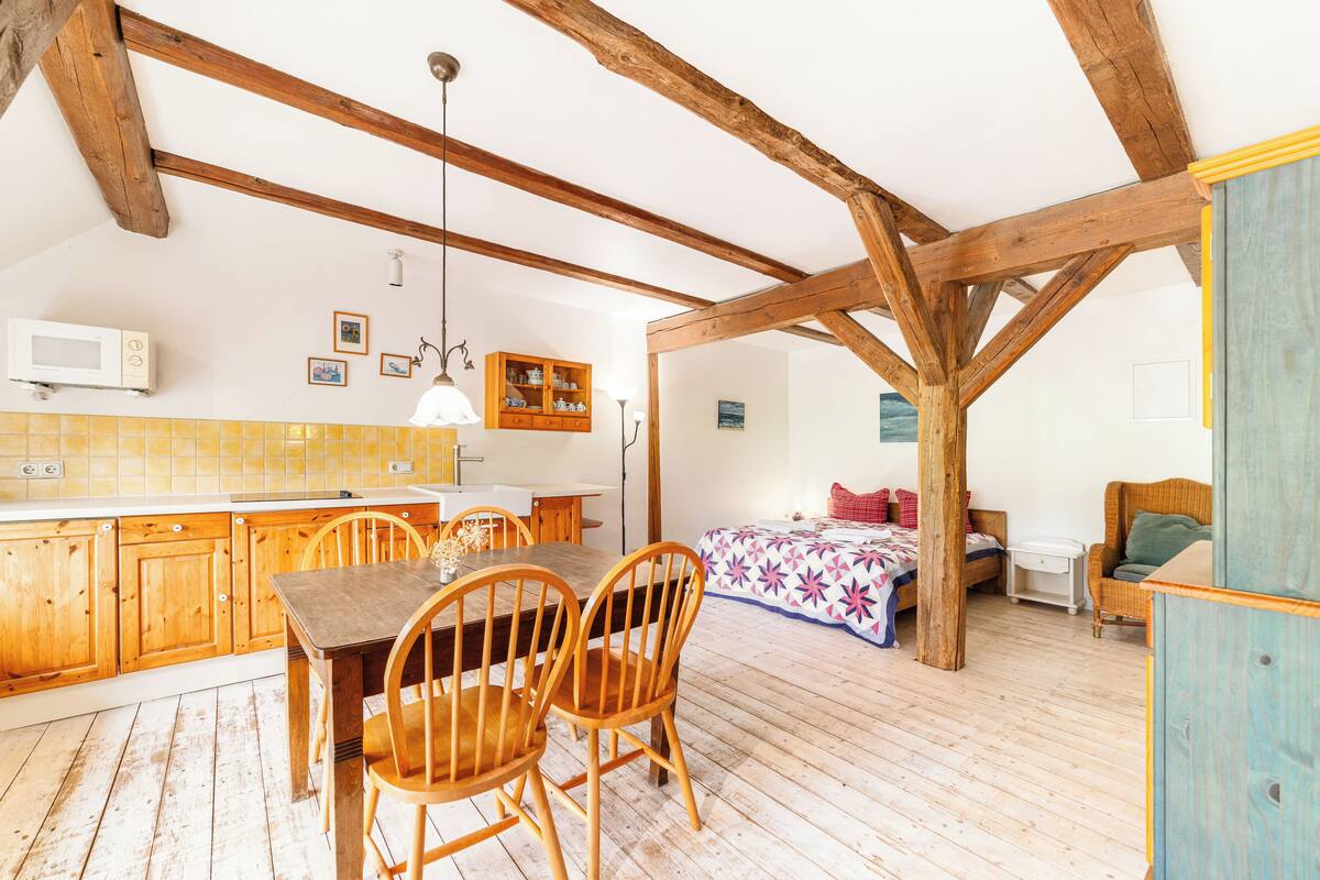 A rustic kitchen with wooden beams, a dining table with chairs, a bed with a colorful bedspread, and a wicker chair.