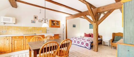 Une cuisine rustique avec des poutres en bois, une table à manger avec des chaises, un lit recouvert d’une courtepointe colorée et un fauteuil en osier.