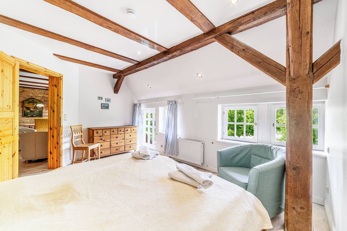 A spacious bedroom with a large bed, wooden beams, a dresser, a chair, and a window with a view of greenery.