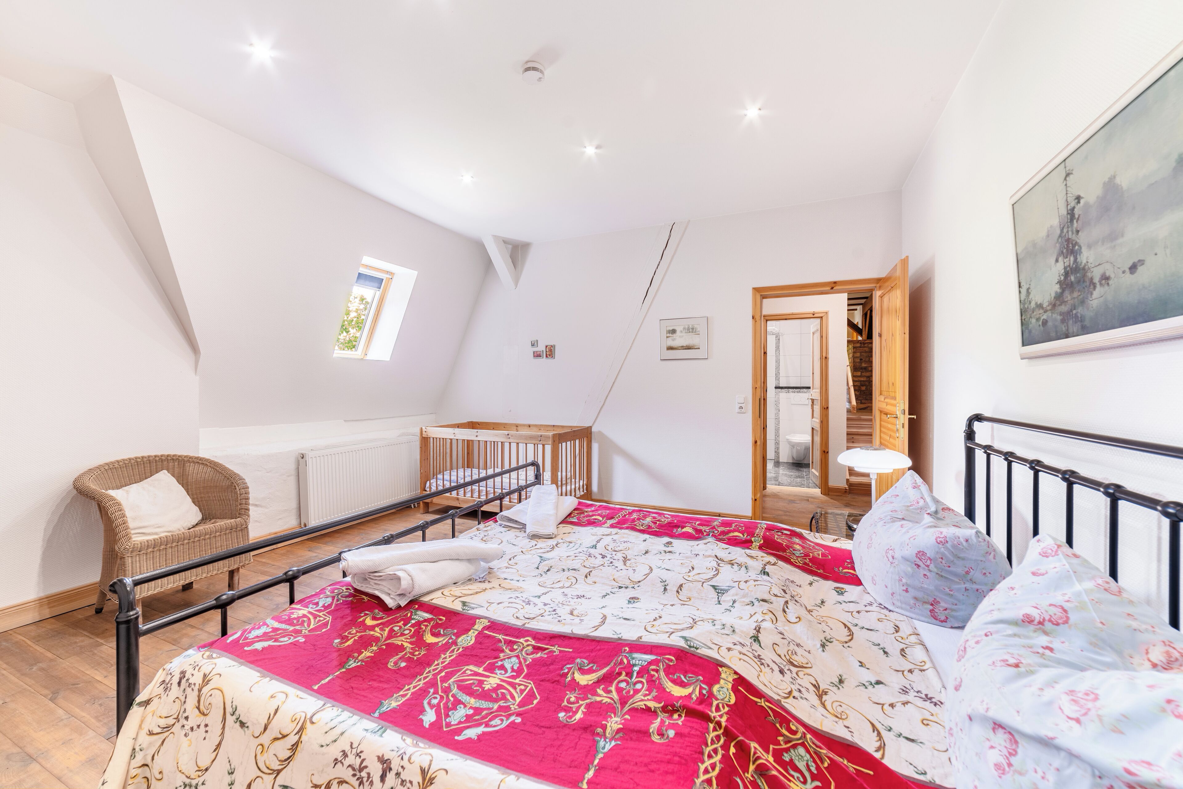 A bedroom with a bed, a chair, a crib, a radiator, a painting, and a door leading to another room.