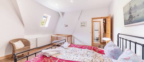 A bedroom with a bed, a chair, a crib, a radiator, a painting, and a door leading to another room.