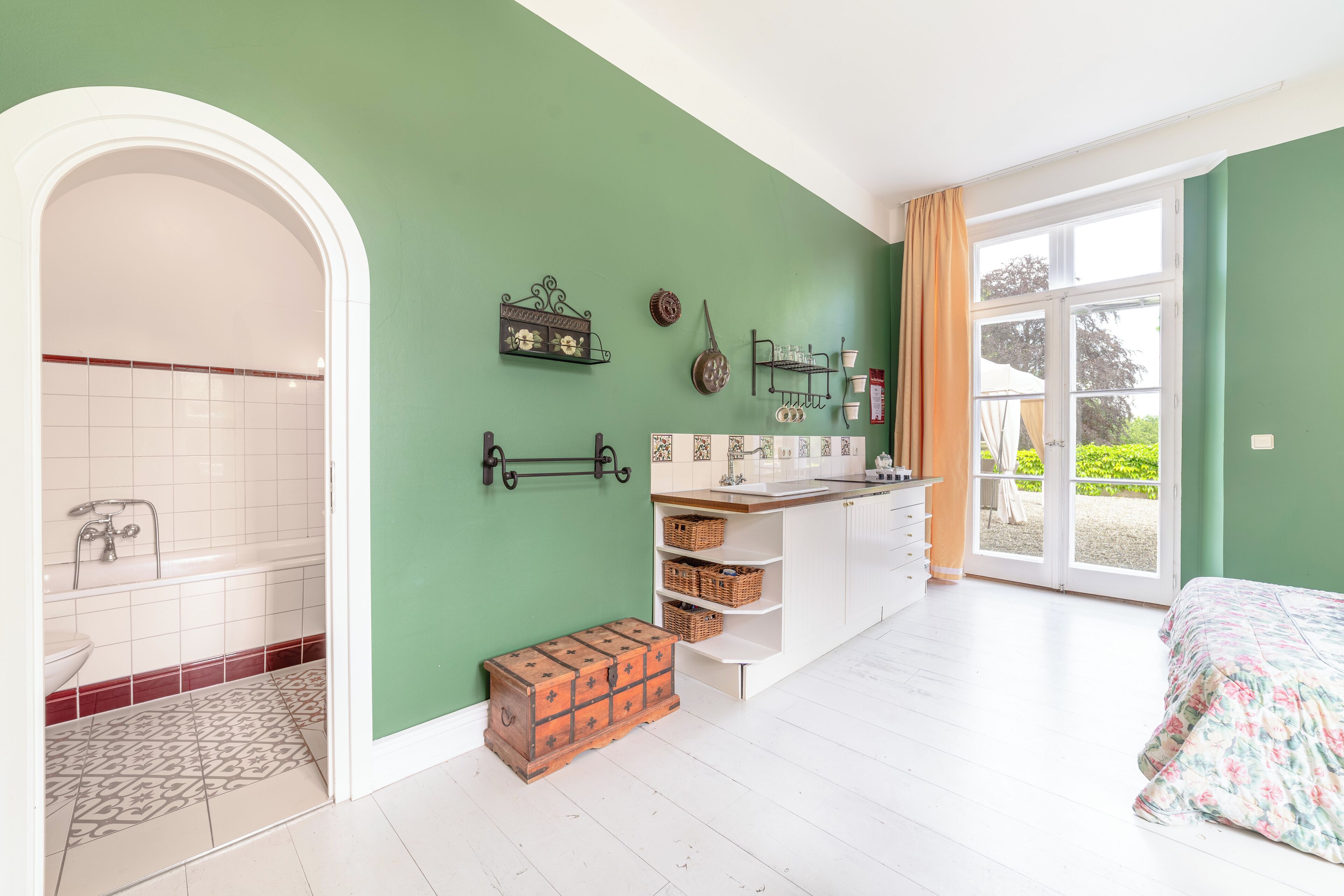 A room with a green wall, a white bathtub, a wooden chest, a bed with floral bedding, and a sliding glass door to the outside.