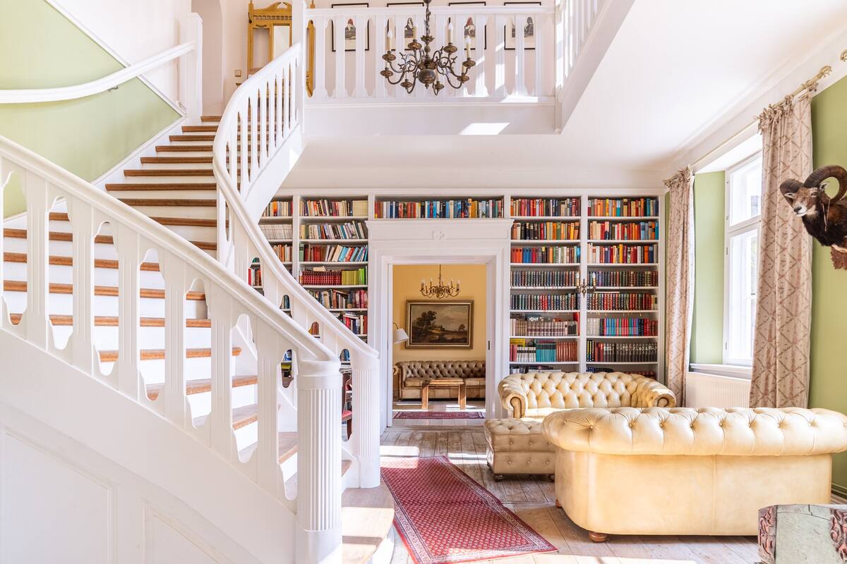 A spacious living area with a staircase leading to a second floor, a large bookcase filled with books, a plush sofa, and a chandelier.