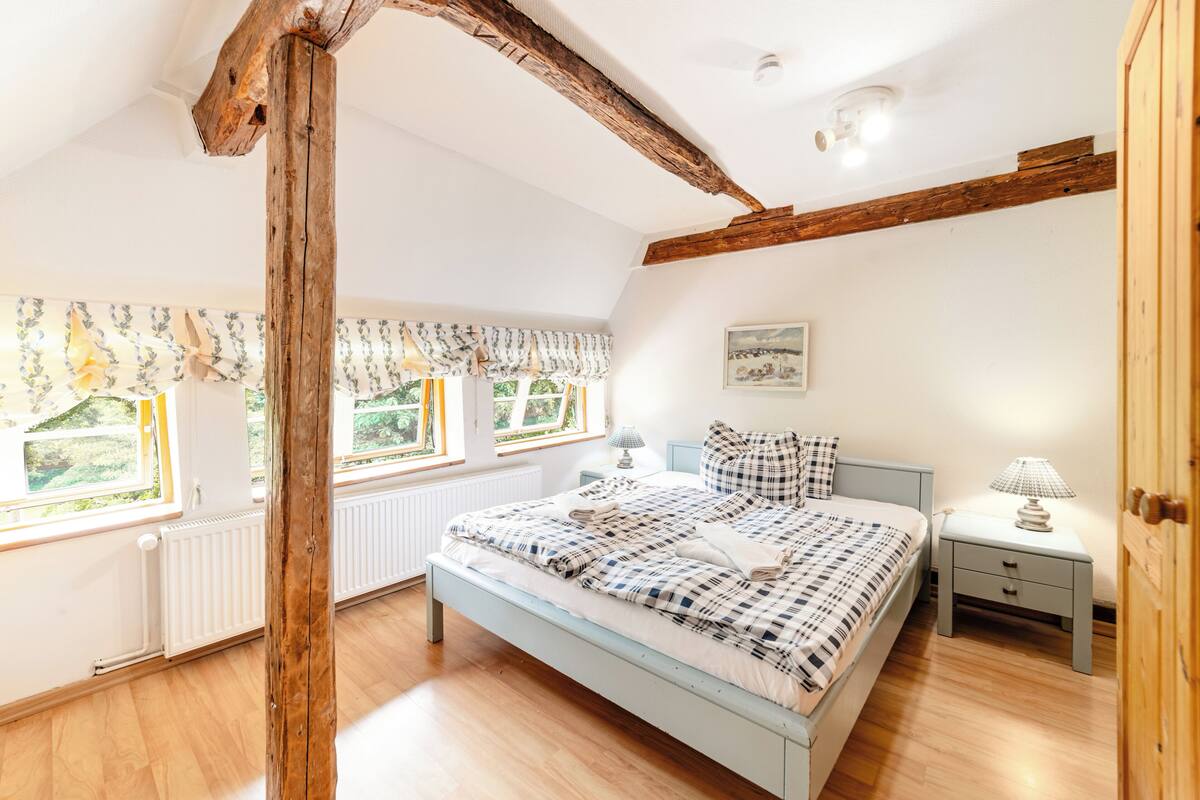 A bedroom with a bed, bedside table, lamp, and windows with curtains.