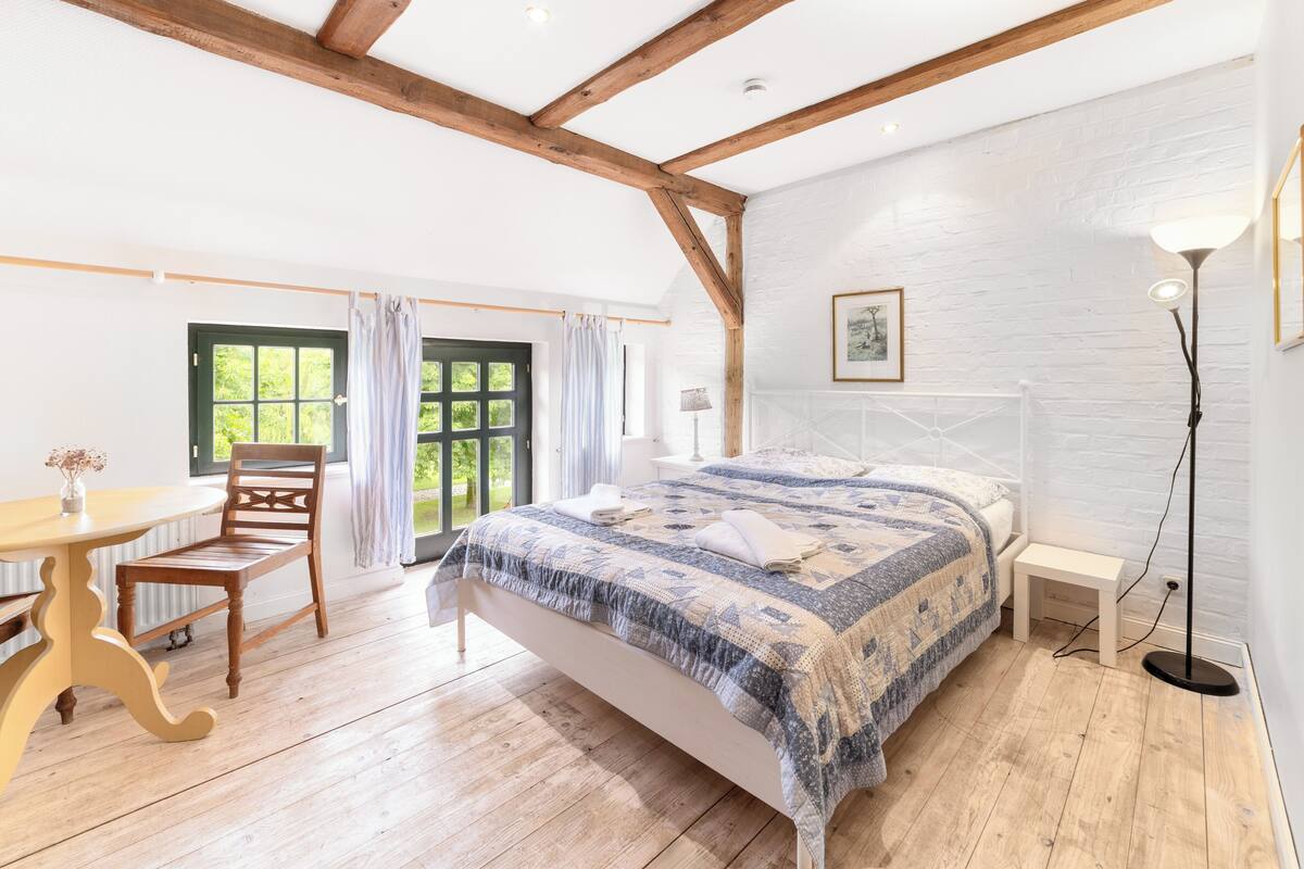 A bedroom with a wooden bed, a small bedside table, a floor lamp, and a window with curtains.